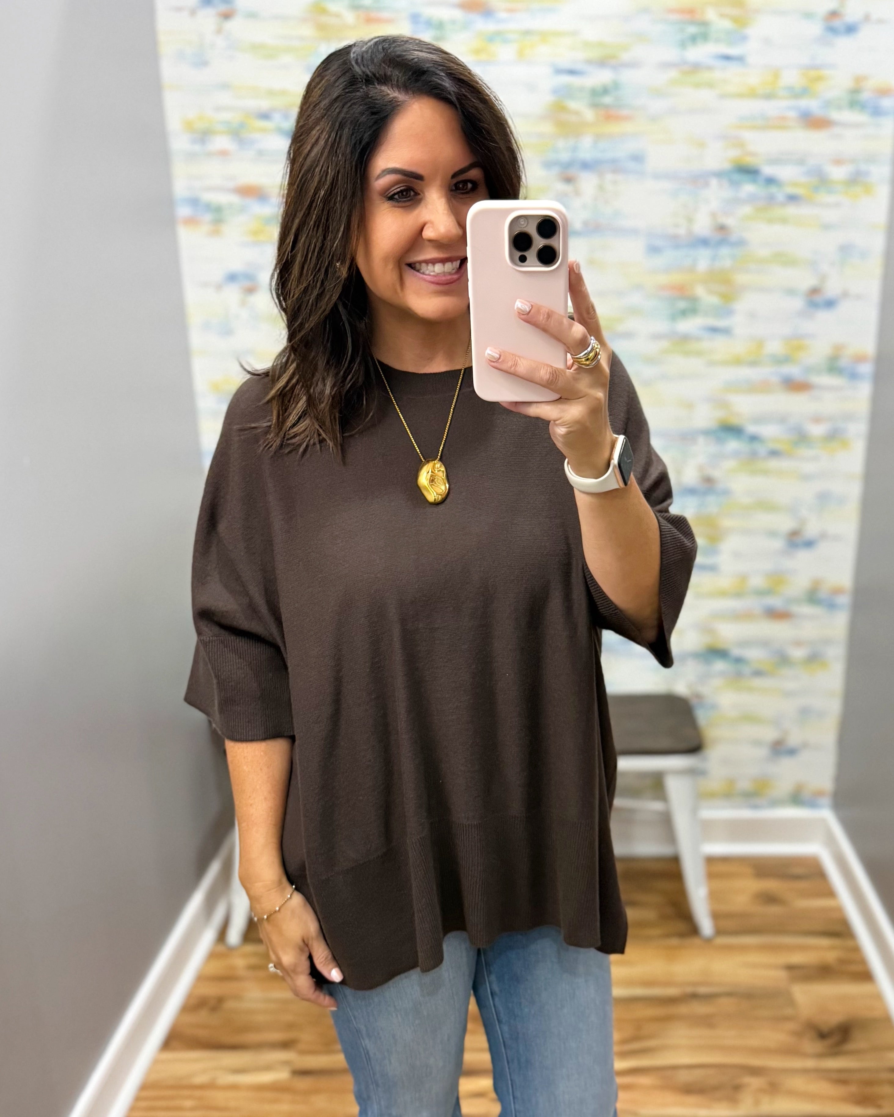 A woman taking a mirror selfie wearing a dark brown, loose-fitting short-sleeve top and light blue jeans in a boutique setting.