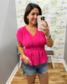 Woman taking a mirror selfie wearing a pink peplum top and denim shorts.