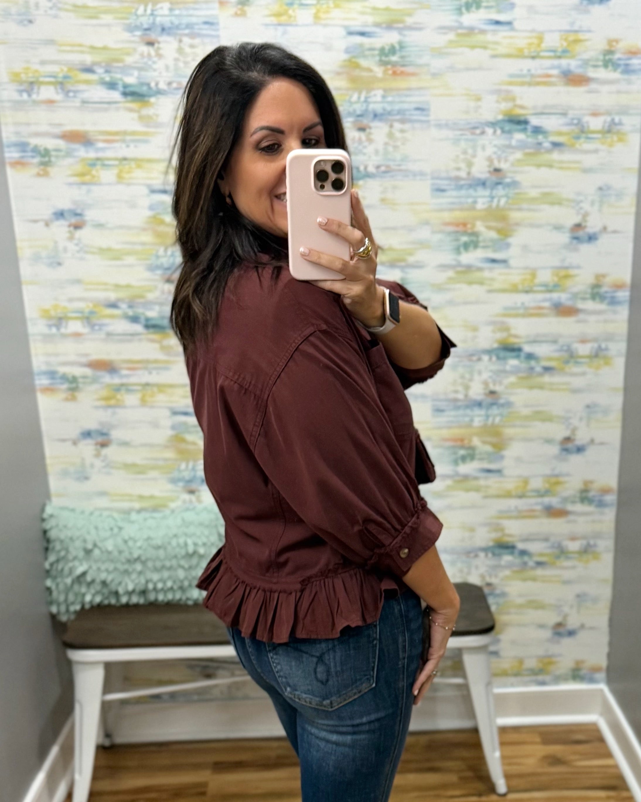 A woman taking a mirror selfie wearing a rust-brown ruffled blouse and blue jeans, standing in front of a patterned wallpaper background.