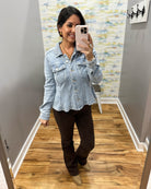 A woman takes a mirror selfie wearing a light-wash blue denim cropped jacket, dark brown pants, and tan boots in a fitting room with wood floors and gray walls.