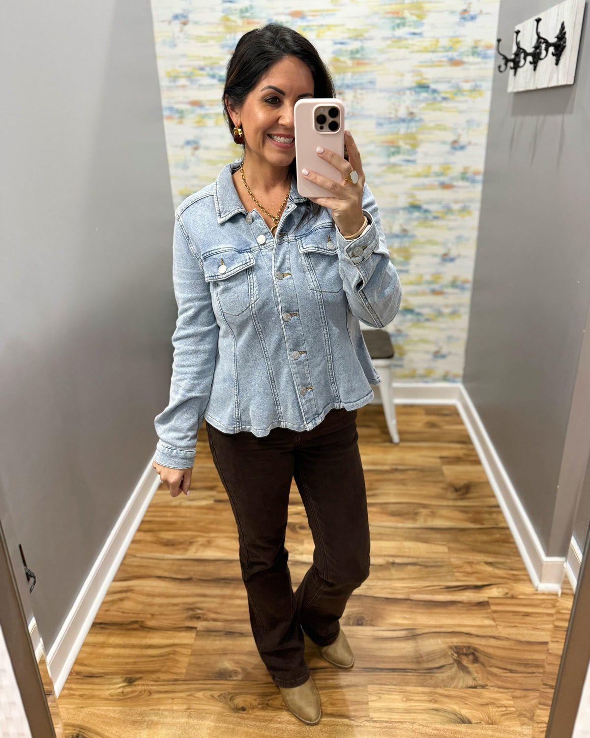 A woman takes a mirror selfie wearing a light-wash blue denim cropped jacket, dark brown pants, and tan boots in a fitting room with wood floors and gray walls.