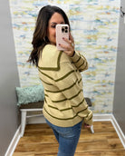A woman takes a mirror selfie in a fitting room wearing a taupe and olive green ribbed knit sweater with olive green horizontal stripes and blue jeans.