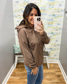 A woman takes a mirror selfie wearing a brown pullover hoodie with a drawstring hood and elastic cuffs, paired with blue jeans in a fitting room with a patterned wall and wood floor.