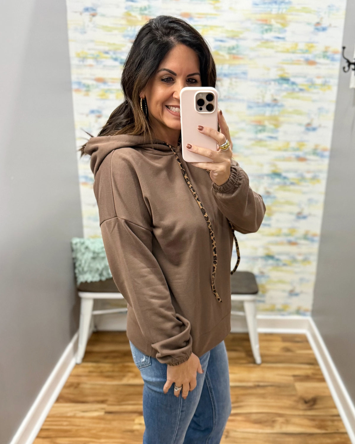 A woman takes a mirror selfie wearing a brown pullover hoodie with a drawstring hood and elastic cuffs, paired with blue jeans in a fitting room with a patterned wall and wood floor.