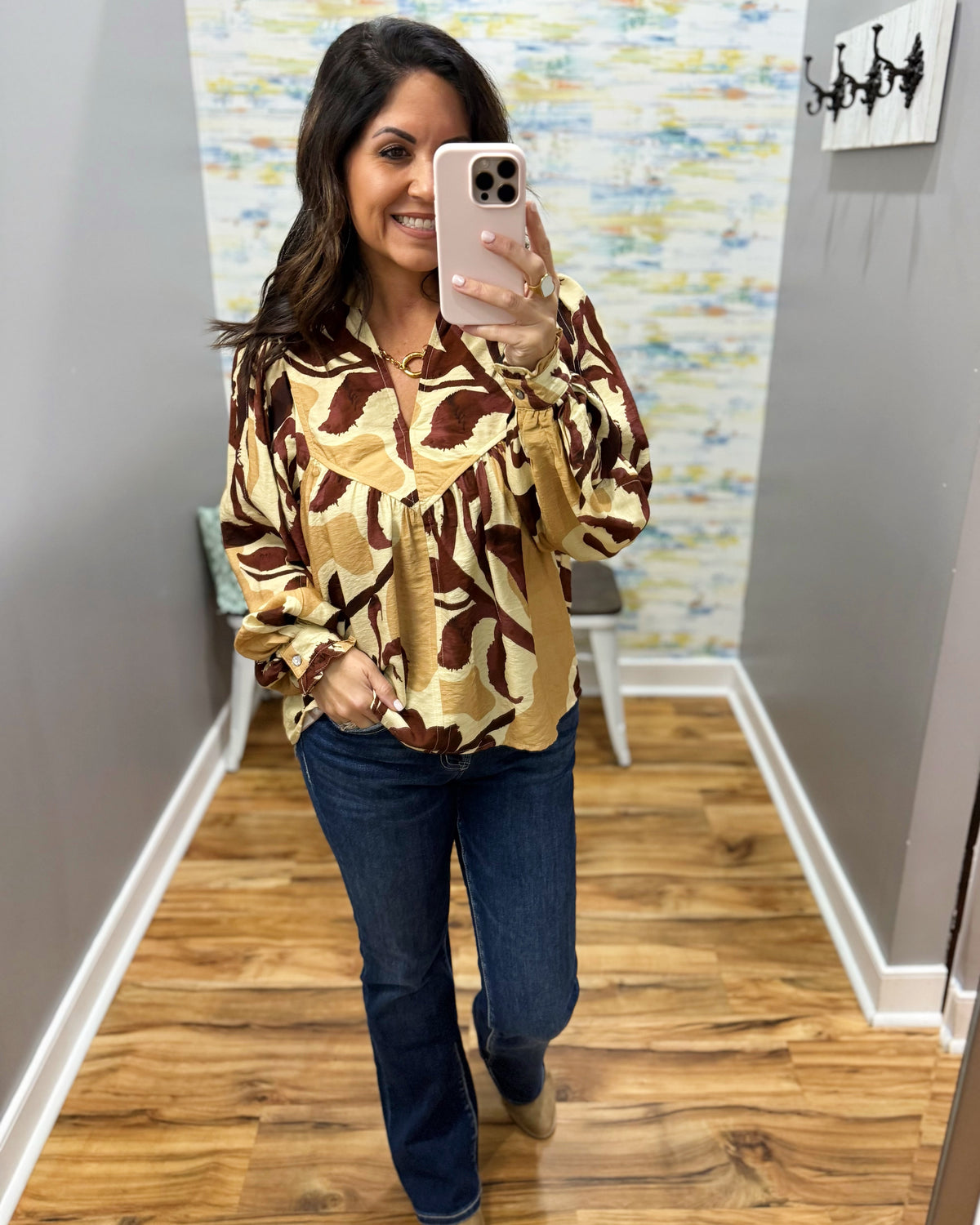 A woman takes a mirror selfie wearing a brown and beige abstract patterned blouse with long sleeves and dark blue jeans, standing in a home hallway.