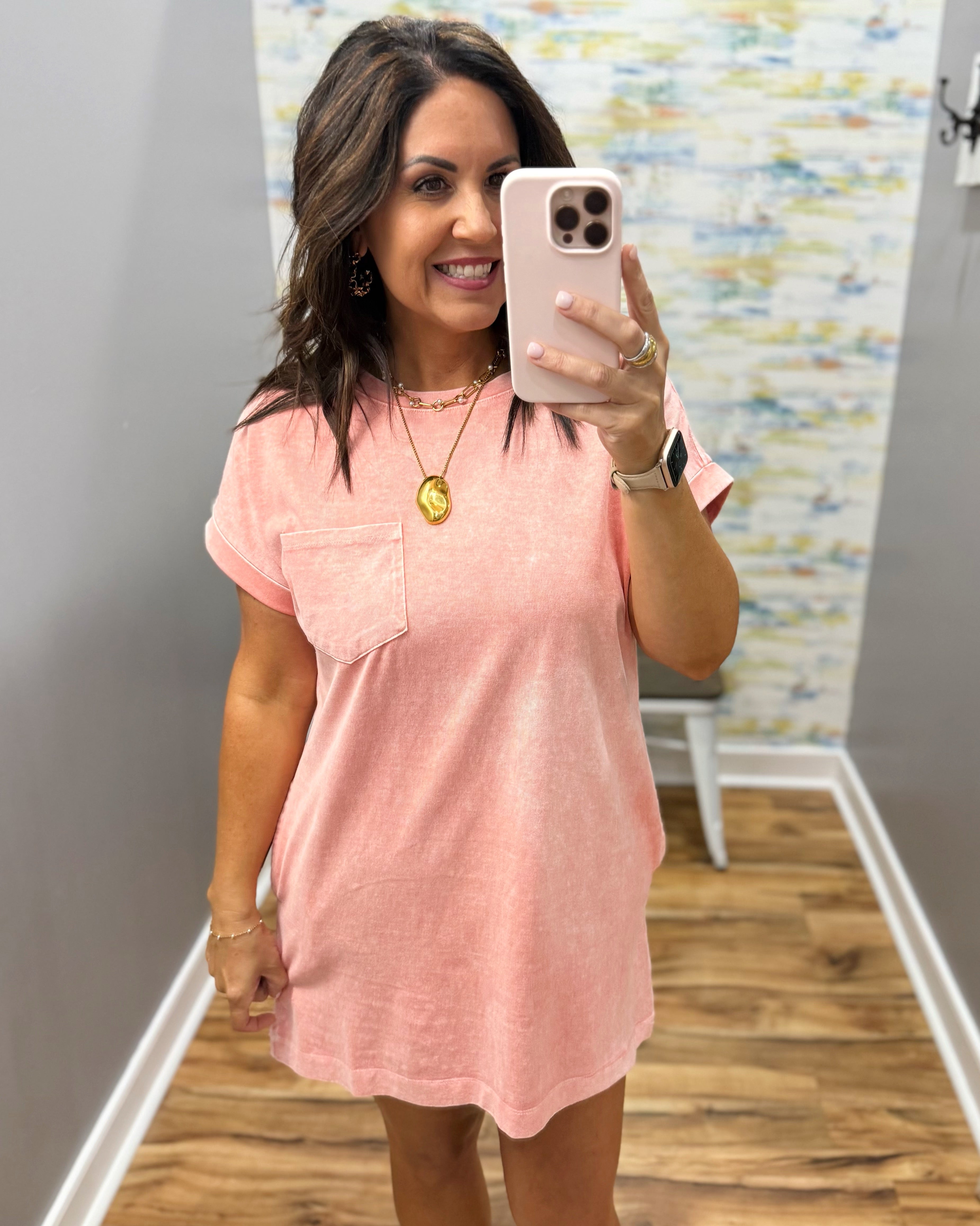 Woman taking a mirror selfie wearing a pink t-shirt dress in a room with a colorful wall.