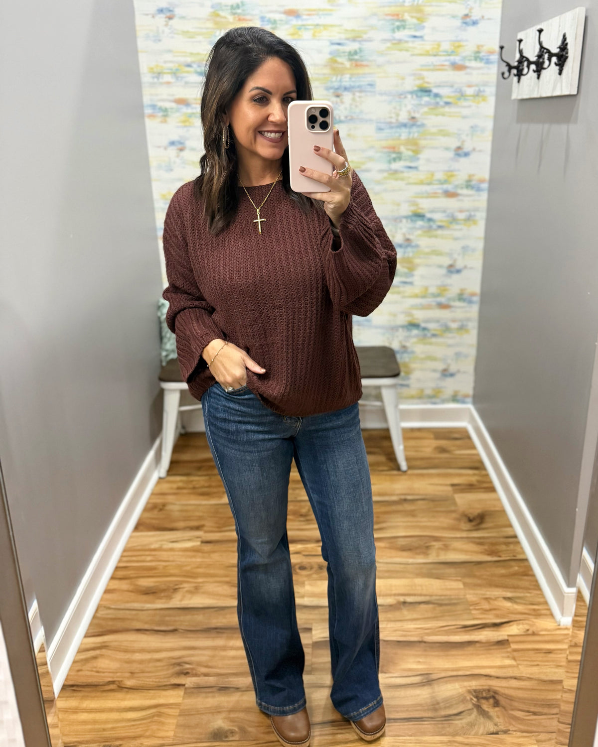 A woman takes a mirror selfie in a fitting room wearing a brown ribbed knit sweater, blue jeans, and brown boots.