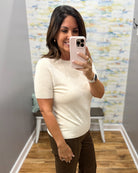 A woman takes a mirror selfie wearing a short-sleeve cream knit top and dark brown pants in a fitting room with a wood floor and abstract wallpaper.