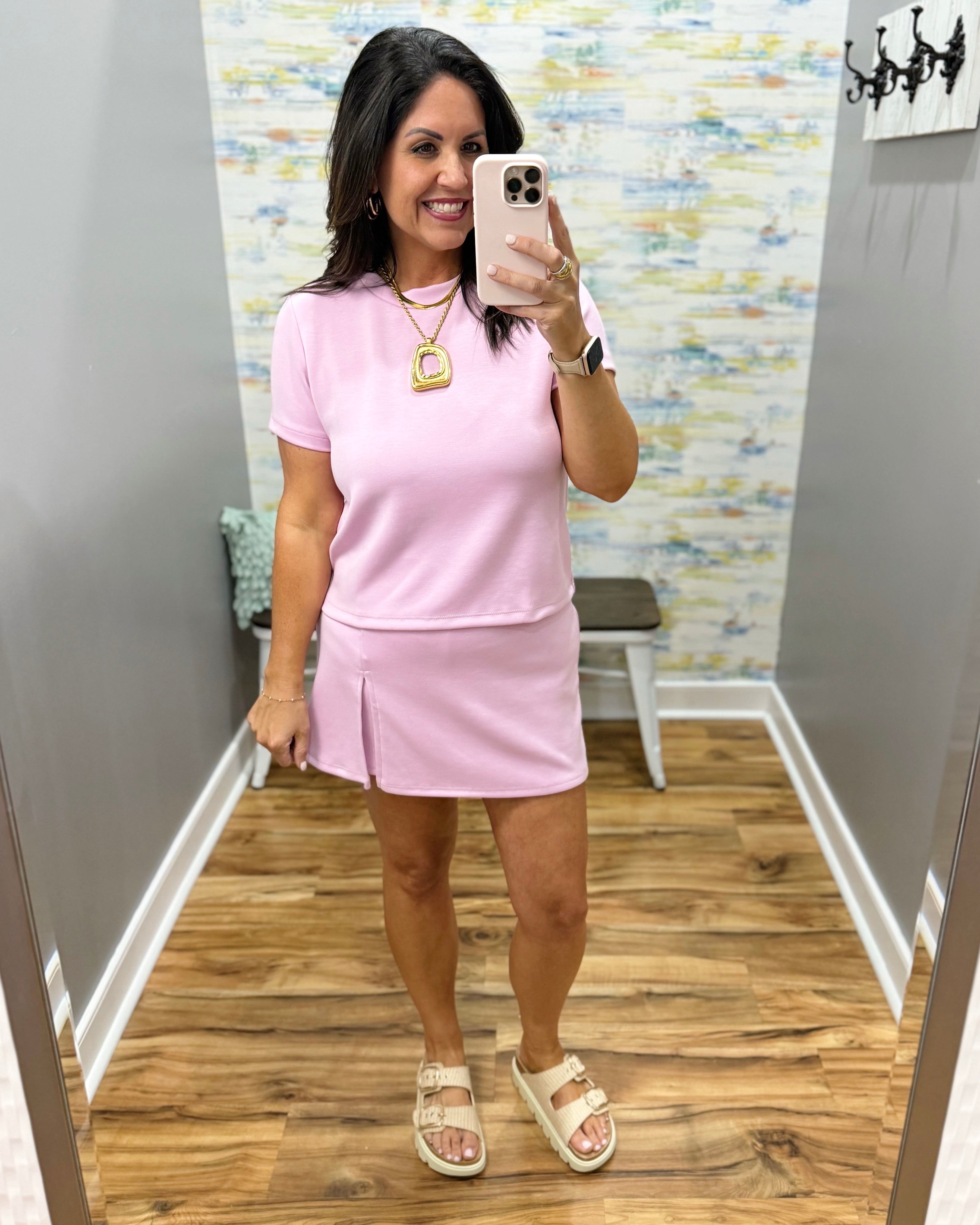 Woman taking a mirror selfie wearing a pink outfit in a room with a patterned wall.