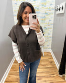 A woman takes a mirror selfie wearing a brown button-front cardigan vest over a white and pink striped long-sleeve shirt, paired with blue jeans, in an indoor hallway setting.