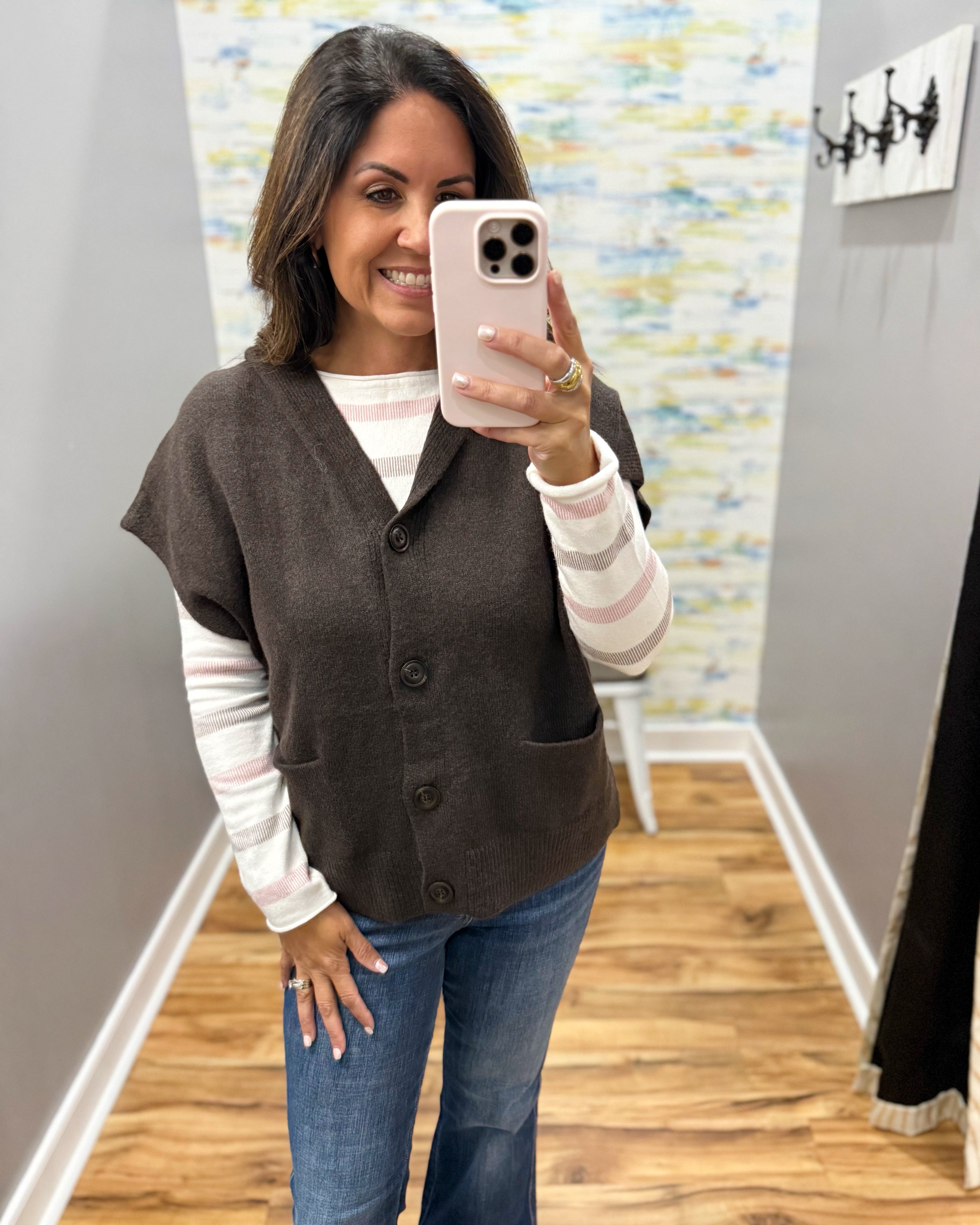 A woman takes a mirror selfie wearing a brown button-front cardigan vest over a white and pink striped long-sleeve shirt, paired with blue jeans, in an indoor hallway setting.
