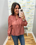 A woman takes a mirror selfie wearing a dusty rose long-sleeve blouse with gathered detailing and blue jeans in a fitting room.