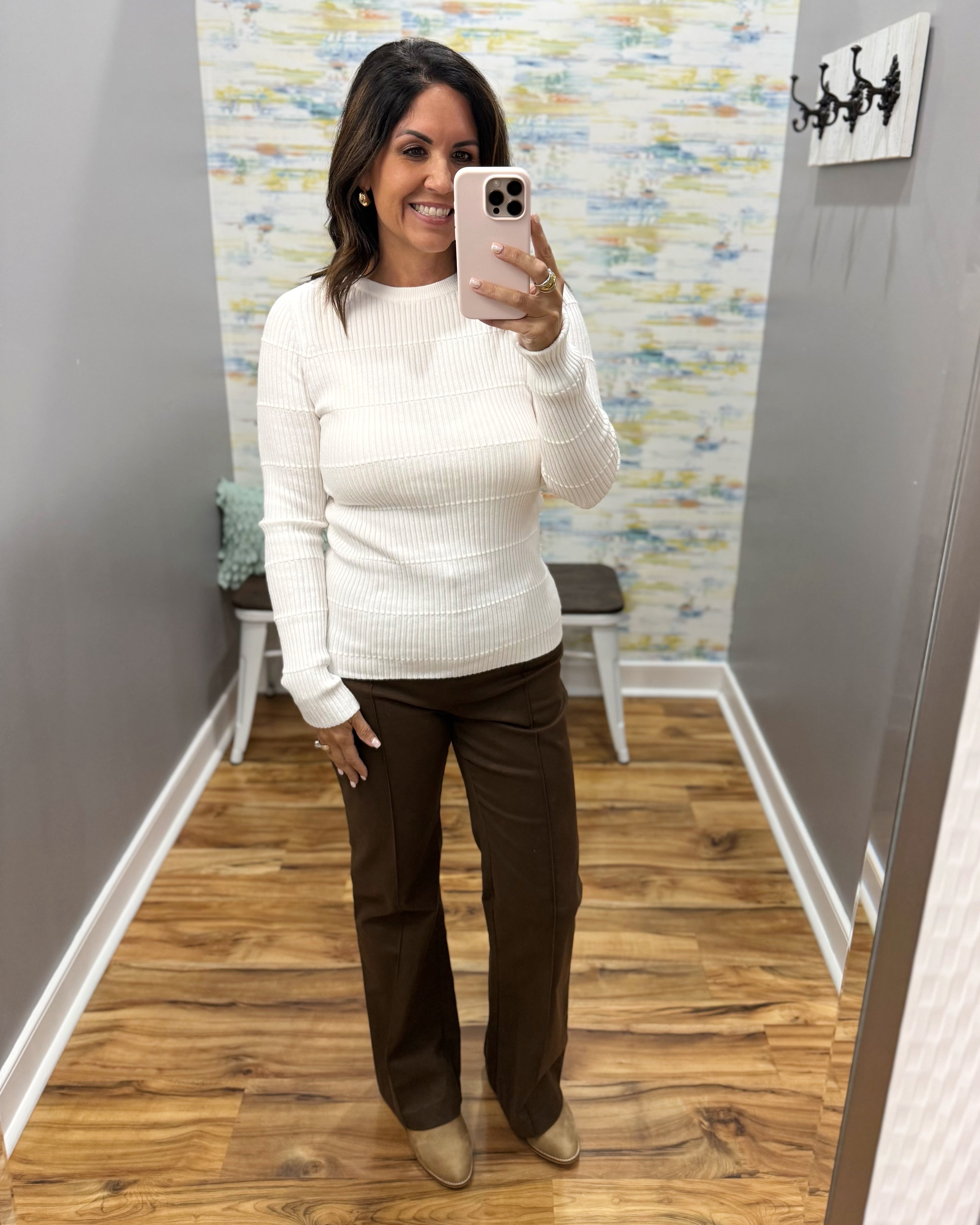A woman takes a mirror selfie wearing a ribbed cream knit sweater and dark brown wide-leg pants in a home setting with wood floors and a patterned wallpaper background.
