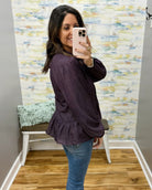 A woman takes a mirror selfie in a fitting room wearing a dark purple long-sleeve blouse with a small white pattern and blue jeans.