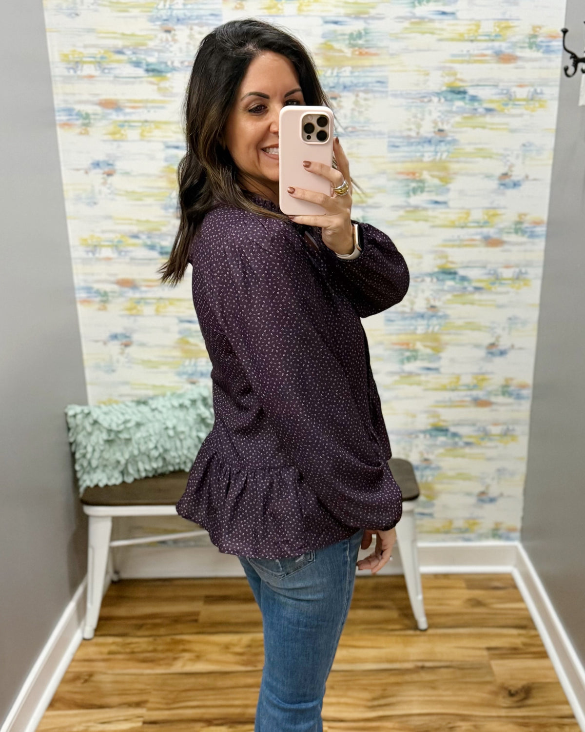 A woman takes a mirror selfie in a fitting room wearing a dark purple long-sleeve blouse with a small white pattern and blue jeans.