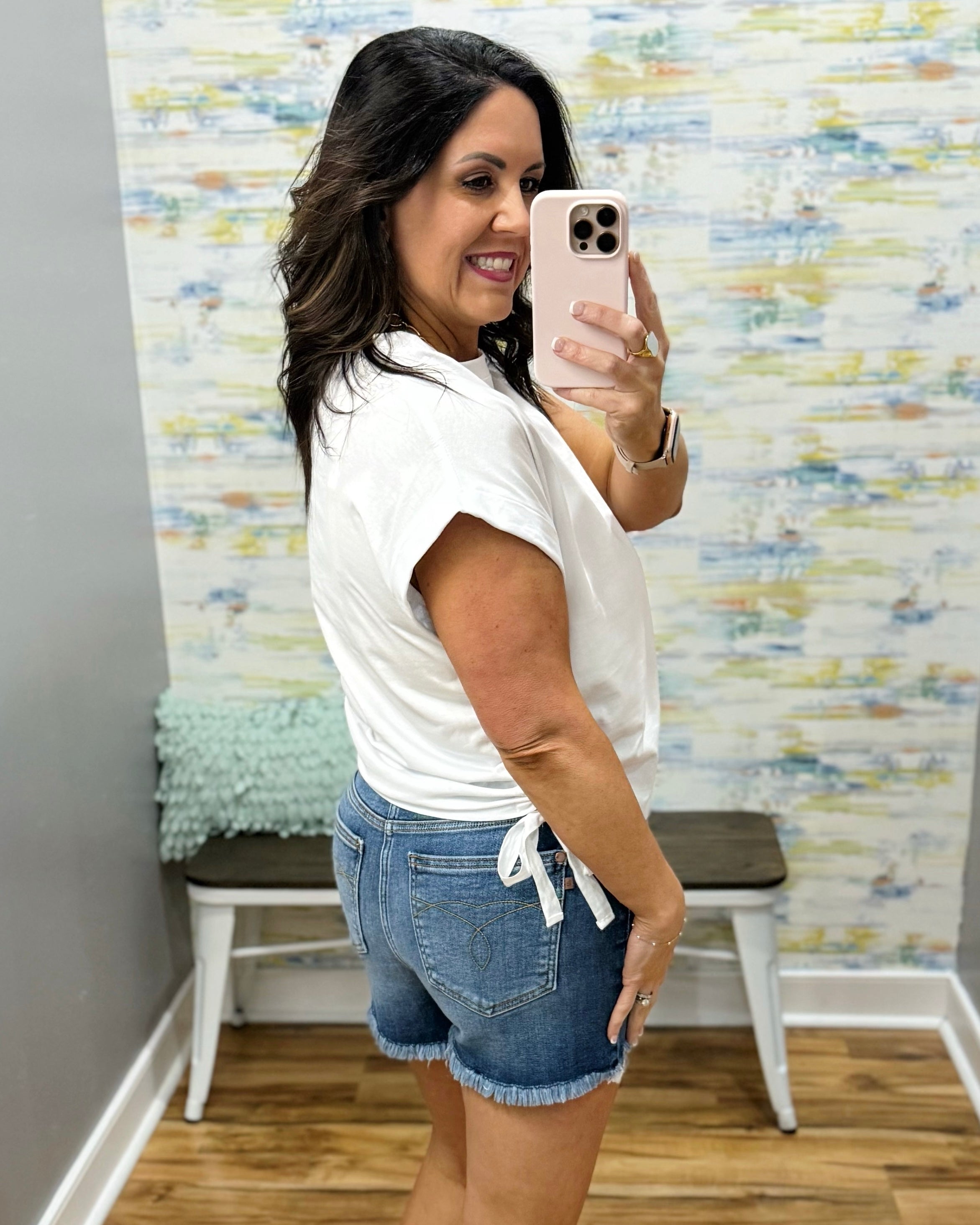 Woman taking a mirror selfie wearing a white top and denim shorts in a room with colorful wall paper.