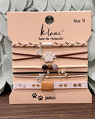 A set of six K'laní hair tie and bracelet pet accessories in Size M, displayed on their original beige cardboard packaging card. The set includes braided, elastic, and beaded options with paw print and K'laní charm details, resting on a dark wooden surface with a gray textured fabric background.