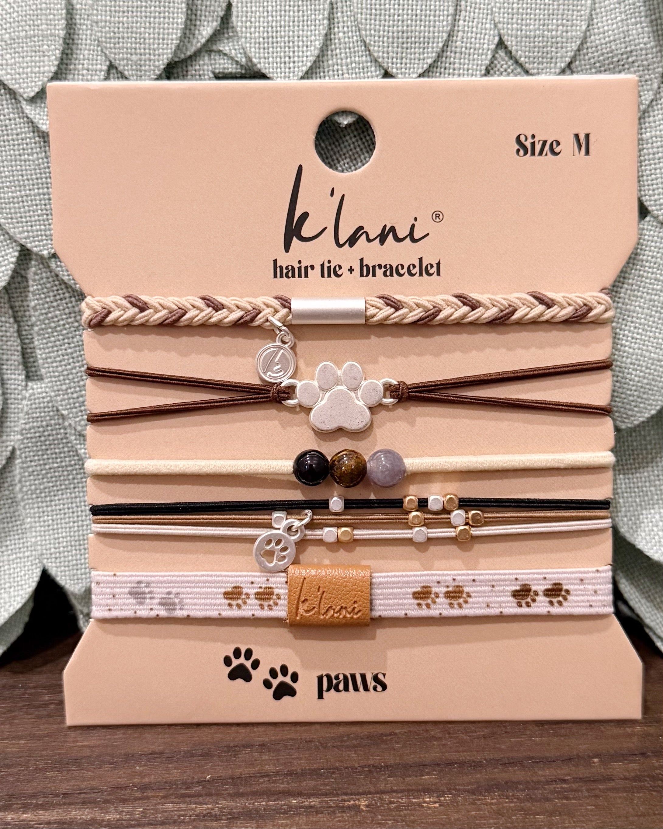 A set of six K'laní hair tie and bracelet pet accessories in Size M, displayed on their original beige cardboard packaging card. The set includes braided, elastic, and beaded options with paw print and K'laní charm details, resting on a dark wooden surface with a gray textured fabric background.