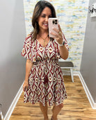 A woman takes a mirror selfie wearing a short-sleeve V-neck dress with a red, beige, and white geometric diamond pattern. She is standing in an indoor hallway with gray walls, wood floors, and a patterned wallpaper background.