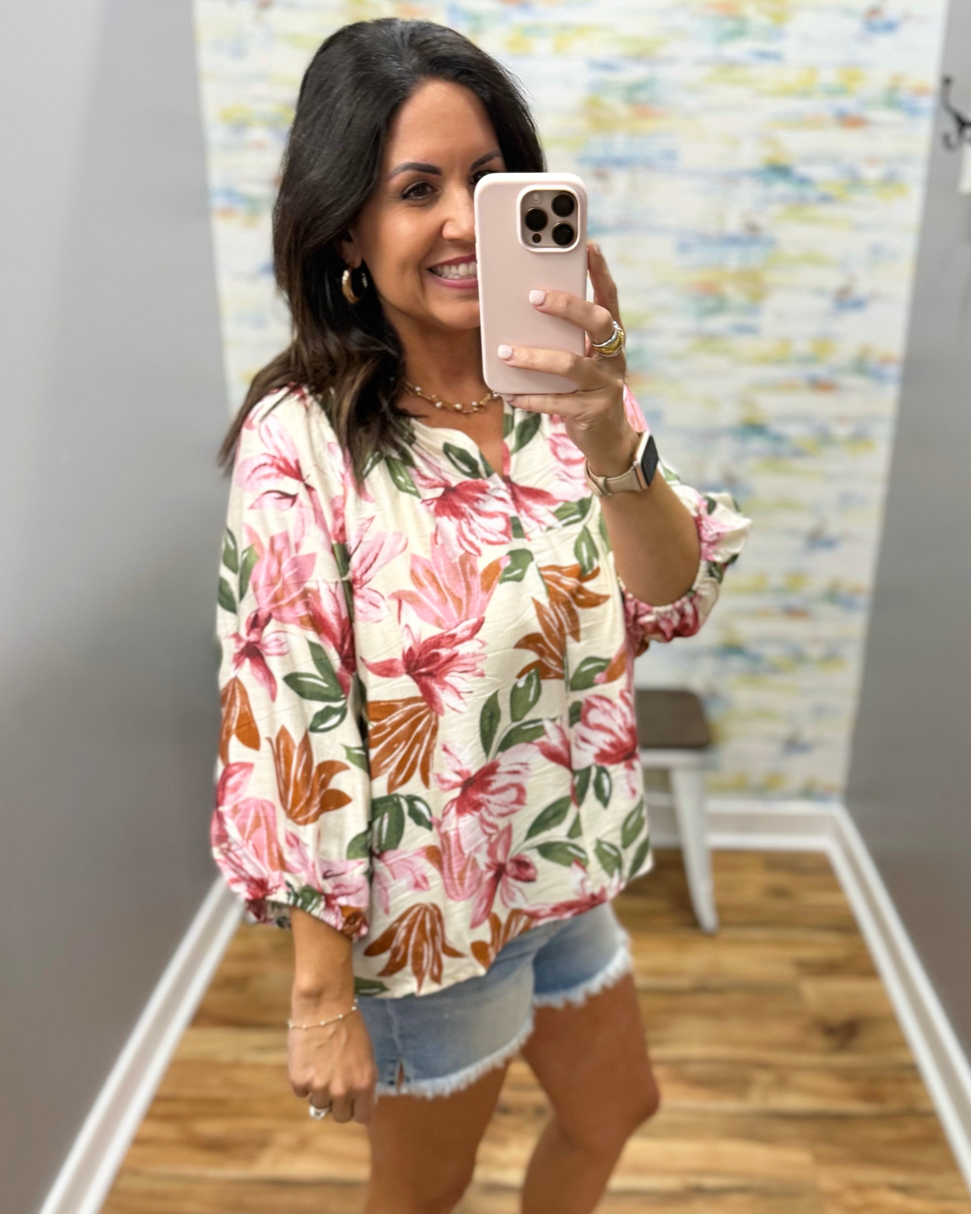 Woman taking a mirror selfie wearing a floral blouse and denim shorts in a room with a colorful wall.