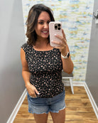 Woman taking a mirror selfie wearing a black floral top and denim shorts in a room with colorful wall art.