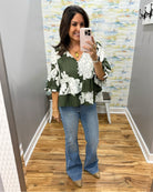 A woman takes a mirror selfie wearing a green and white floral print blouse with tiered ruffle sleeves, paired with blue flare jeans and tan pointed-toe flats, in a fitting room with wood floors.
