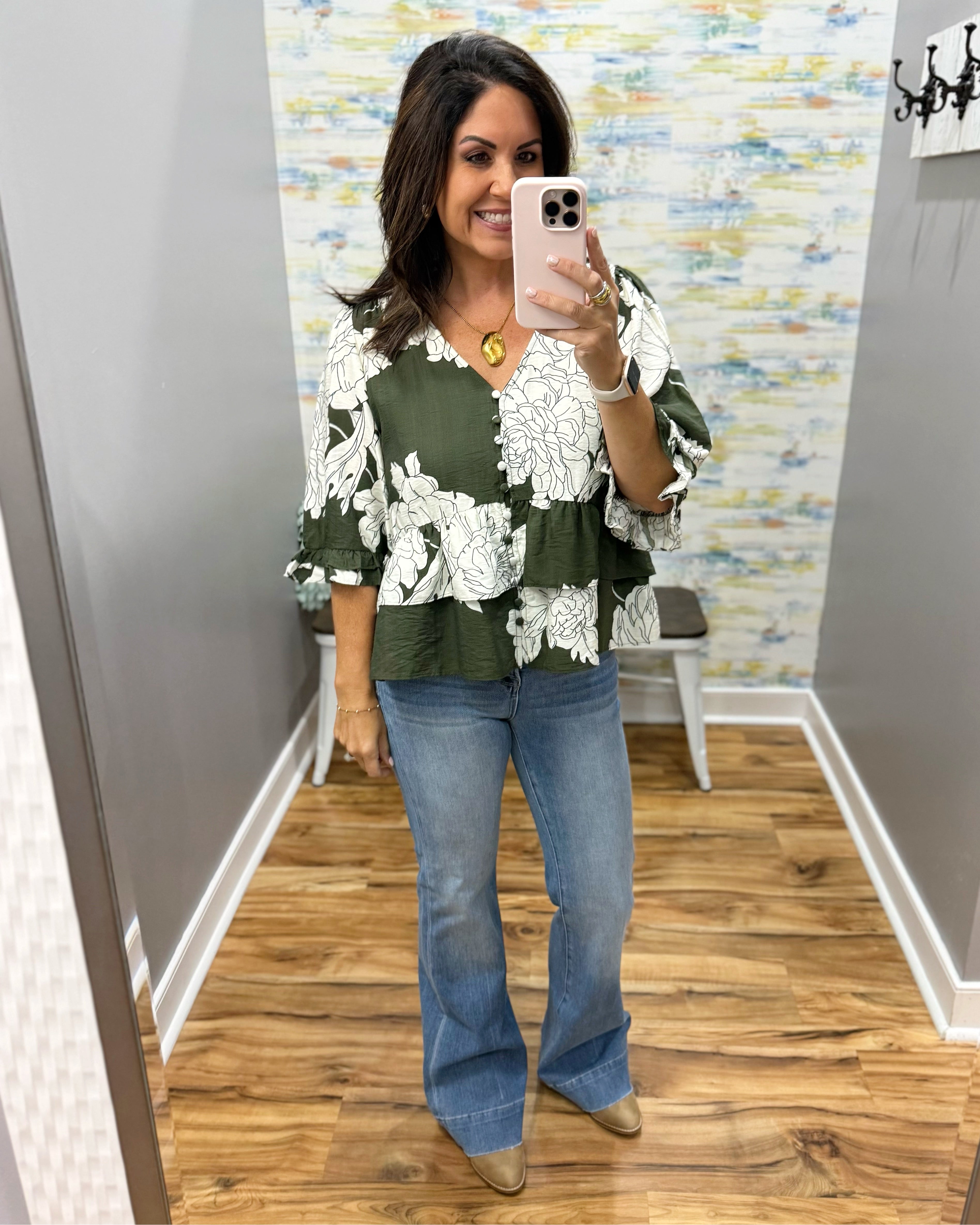 A woman takes a mirror selfie wearing a green and white floral print blouse with tiered ruffle sleeves, paired with blue flare jeans and tan pointed-toe flats, in a fitting room with wood floors.