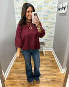 A woman takes a mirror selfie in a fitting room or home interior, wearing a loose burgundy long-sleeve top and blue flare jeans with brown boots. The background shows gray walls, a wood floor, and a decorative abstract wallpaper.