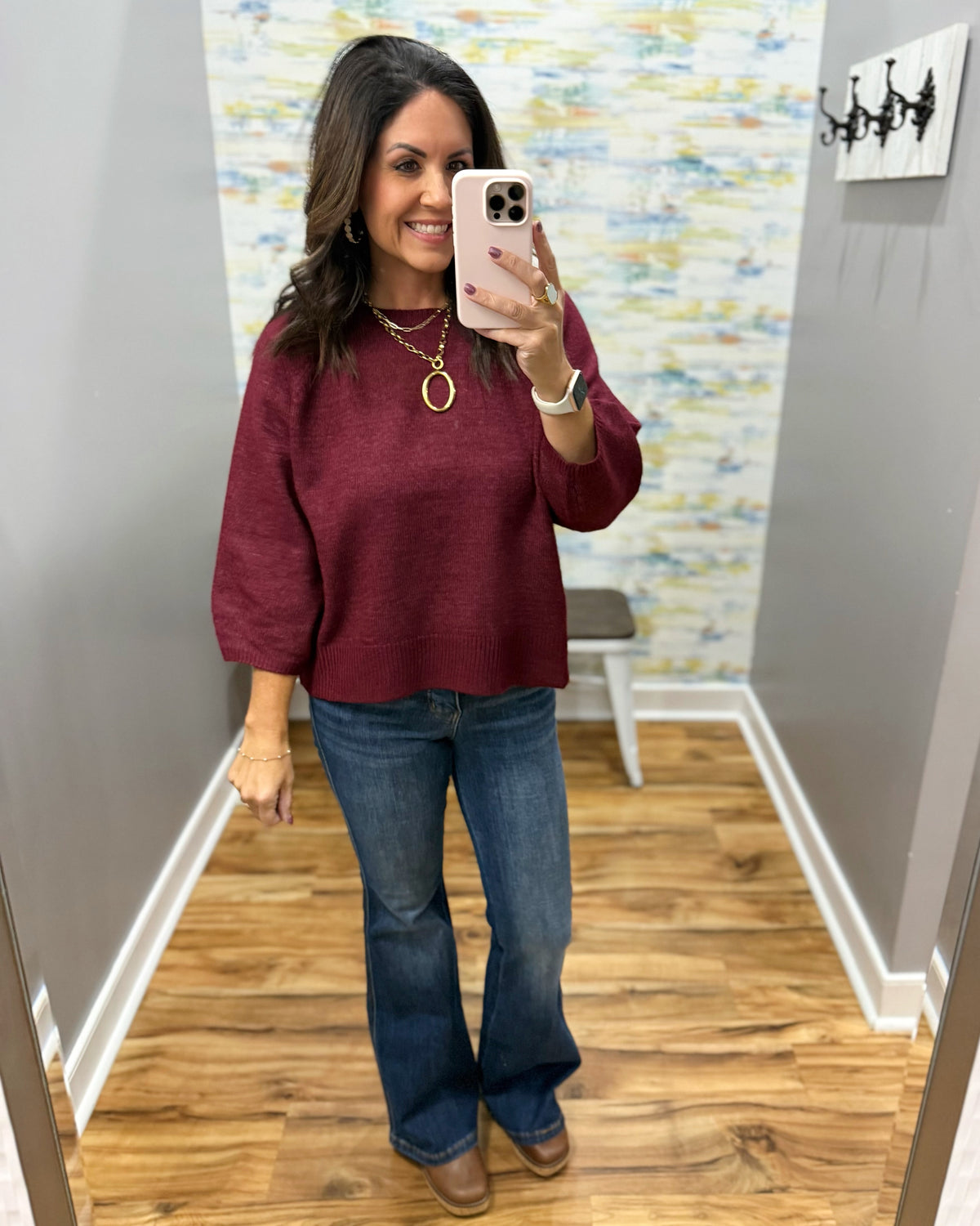 A woman takes a mirror selfie in a fitting room or home interior, wearing a loose burgundy long-sleeve top and blue flare jeans with brown boots. The background shows gray walls, a wood floor, and a decorative abstract wallpaper.