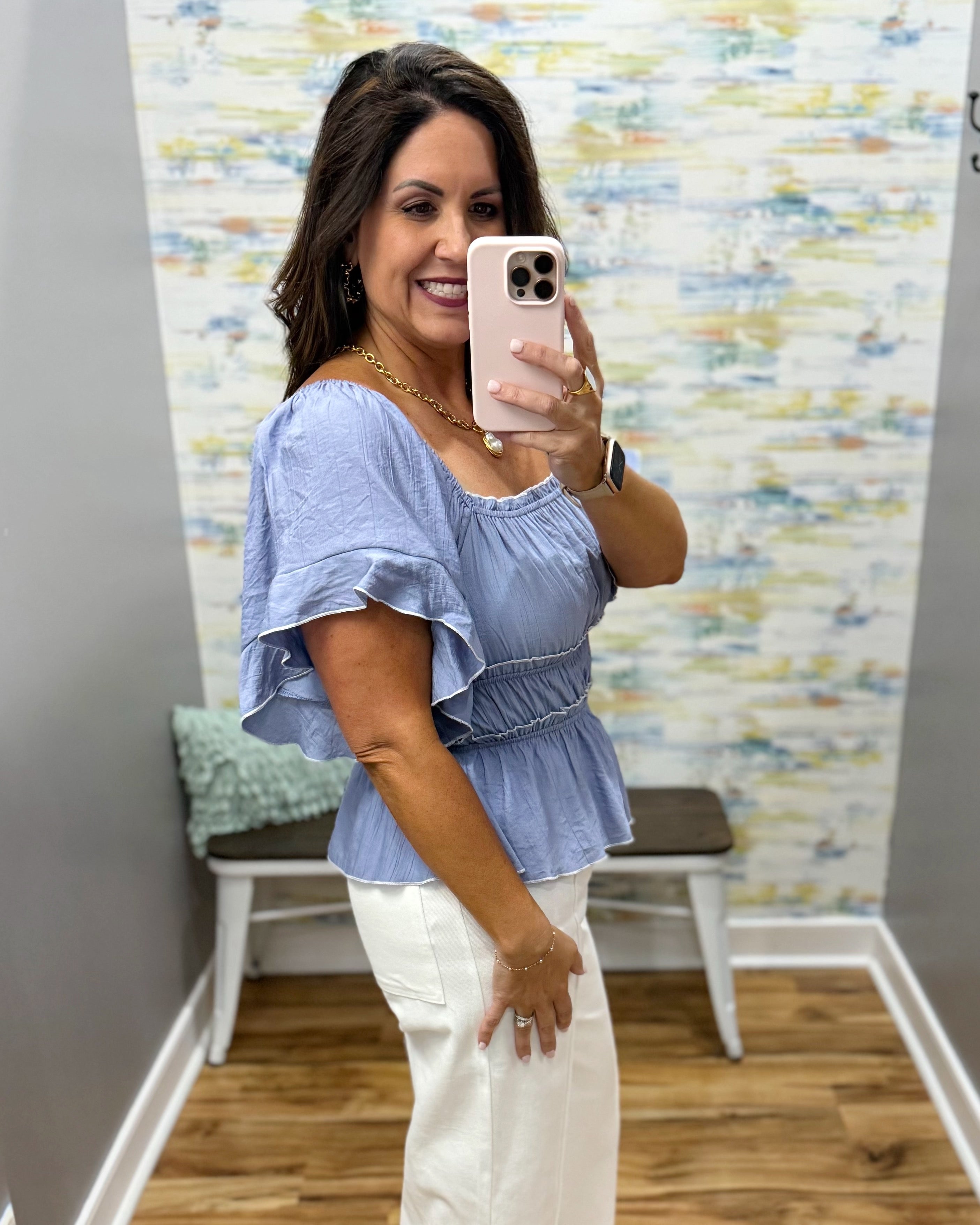 Woman taking a mirror selfie wearing a blue top and white pants in a room with a colorful wall.