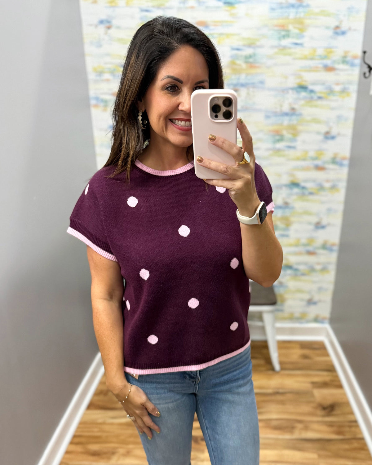A woman taking a mirror selfie in a boutique wearing a short-sleeve knit top in plum with light pink trim and white polka dots, paired with blue jeans.