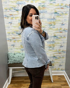 A woman taking a mirror selfie in a fitting room wearing a light-wash denim jacket and dark brown pants, with a patterned wallpaper background.
