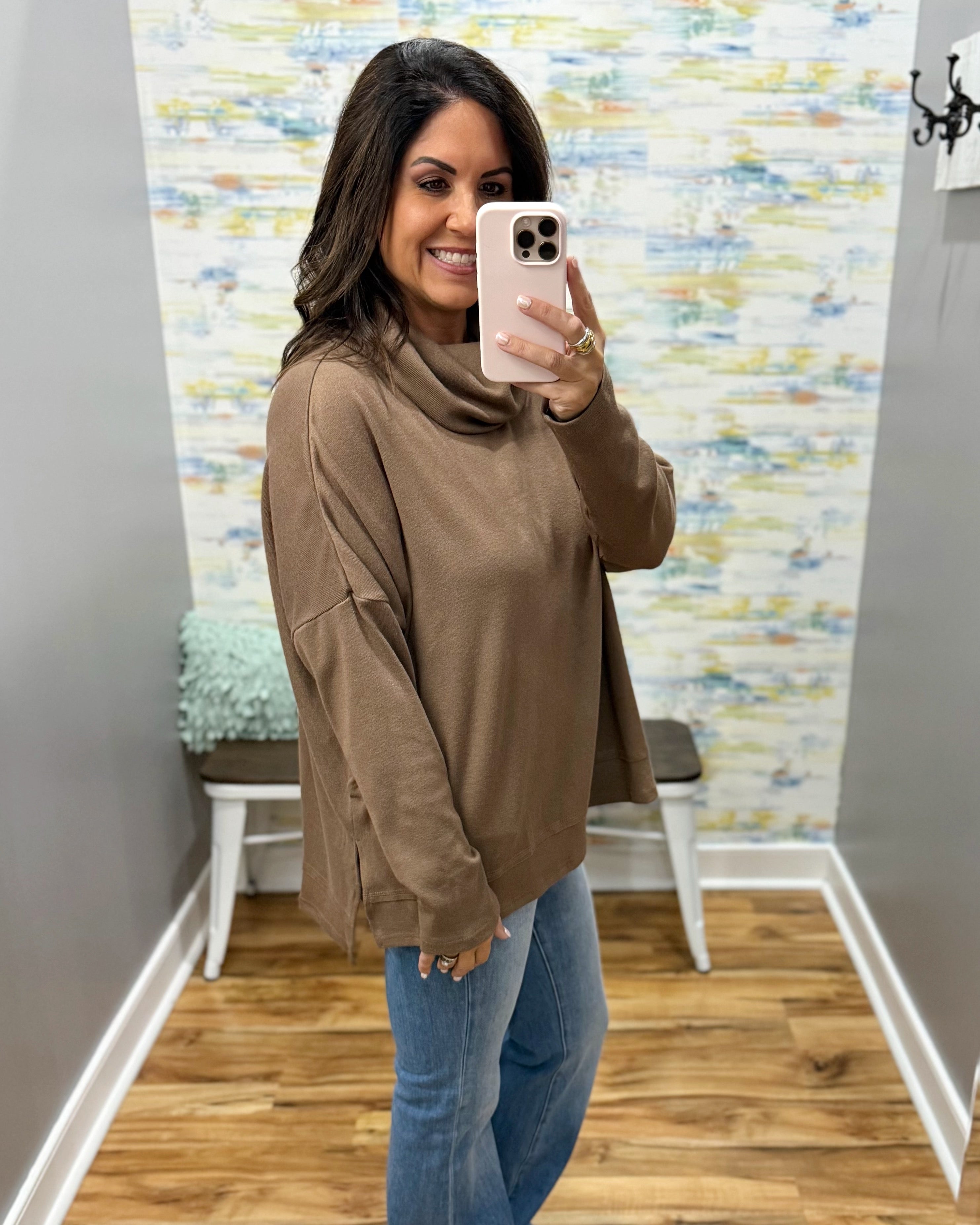 A woman takes a mirror selfie wearing a brown cowl-neck long-sleeve top and blue jeans in a room with a patterned wallpaper background.
