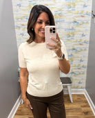 A woman taking a mirror selfie in a fitting room wearing a short-sleeve cream knit top and brown pants.