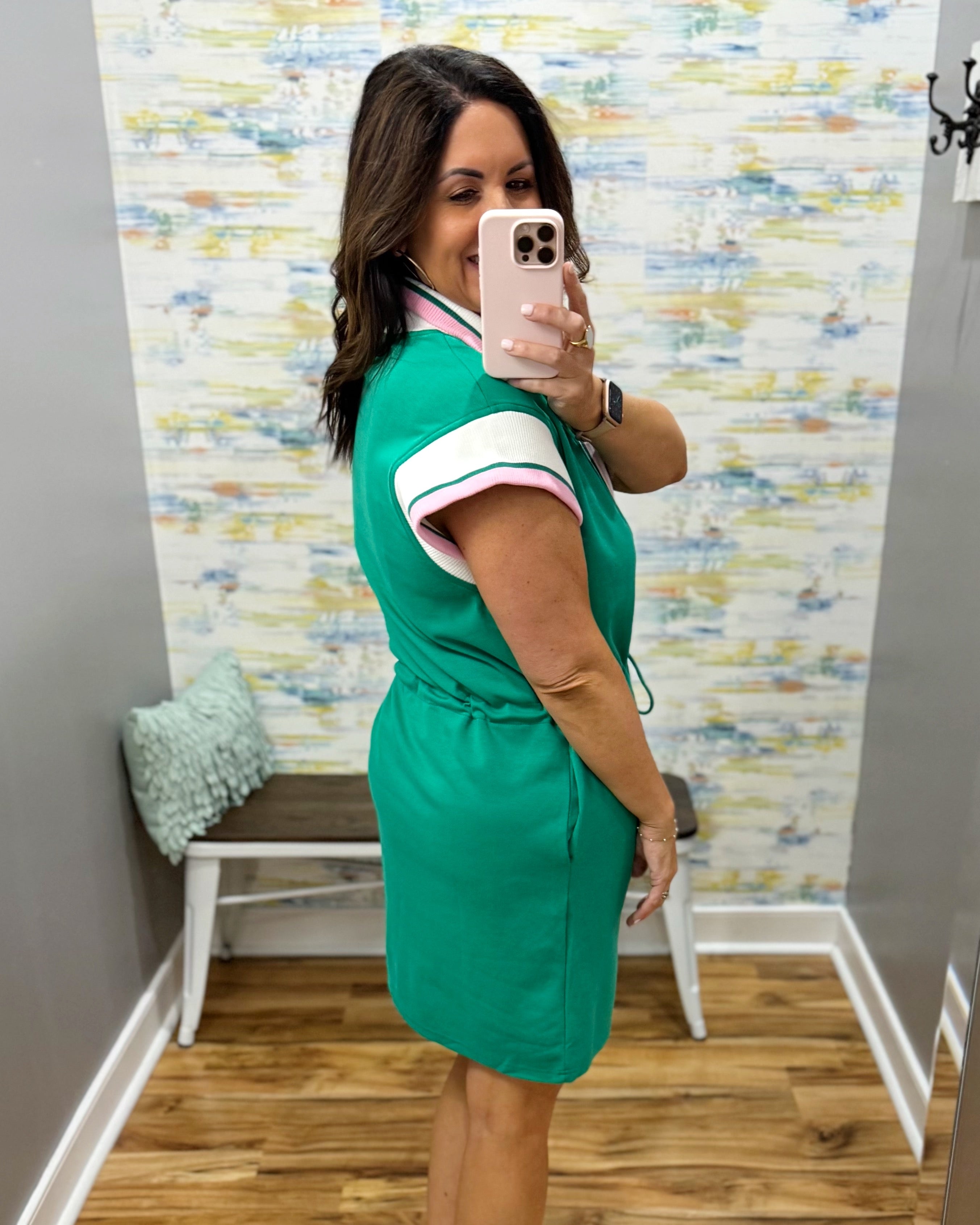 Woman taking a mirror selfie wearing a green dress with a colorful wall and chair in the background.
