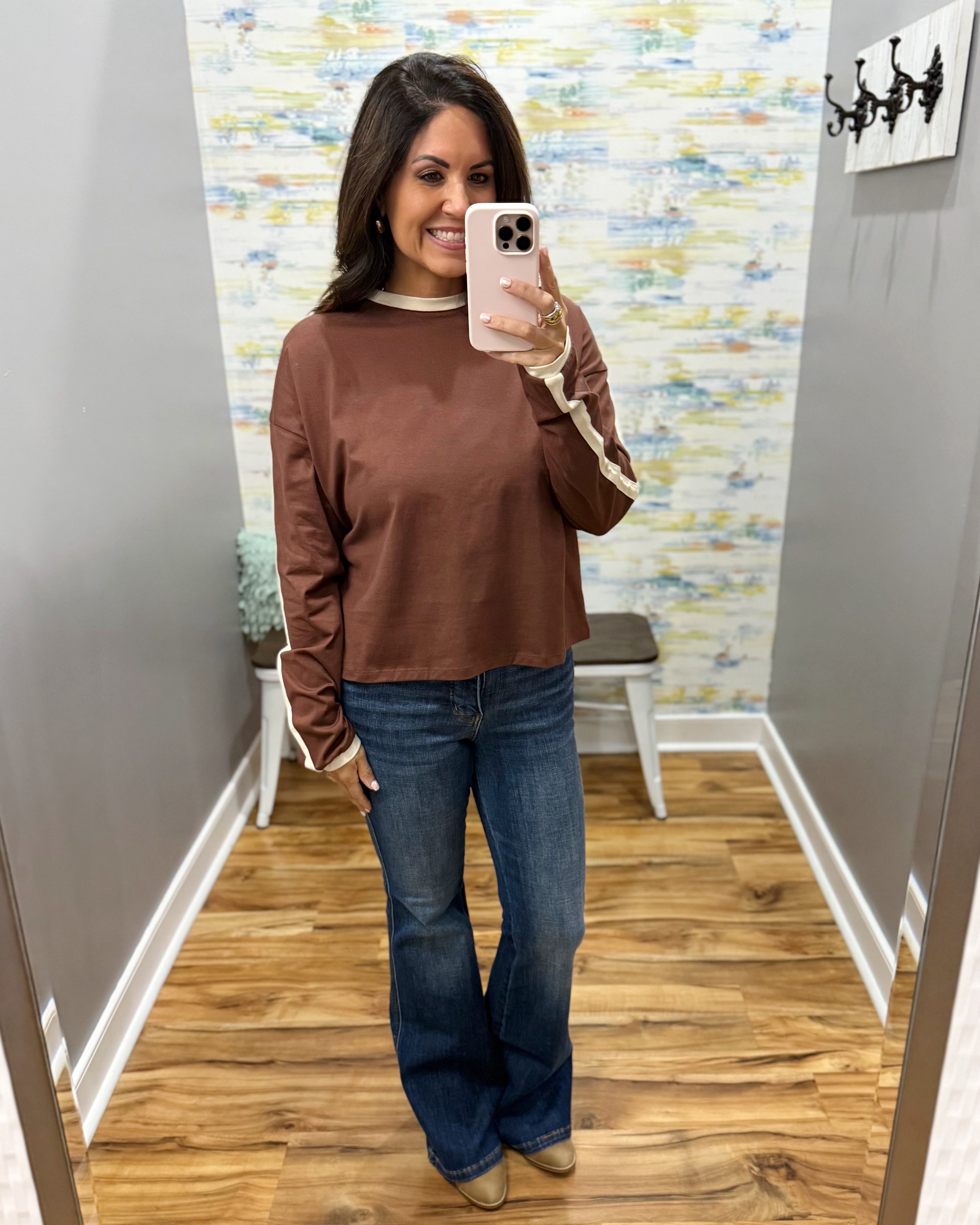 A woman takes a mirror selfie in a fitting room wearing a brown long-sleeve top with beige stripes on the sleeves, blue jeans, and tan shoes.