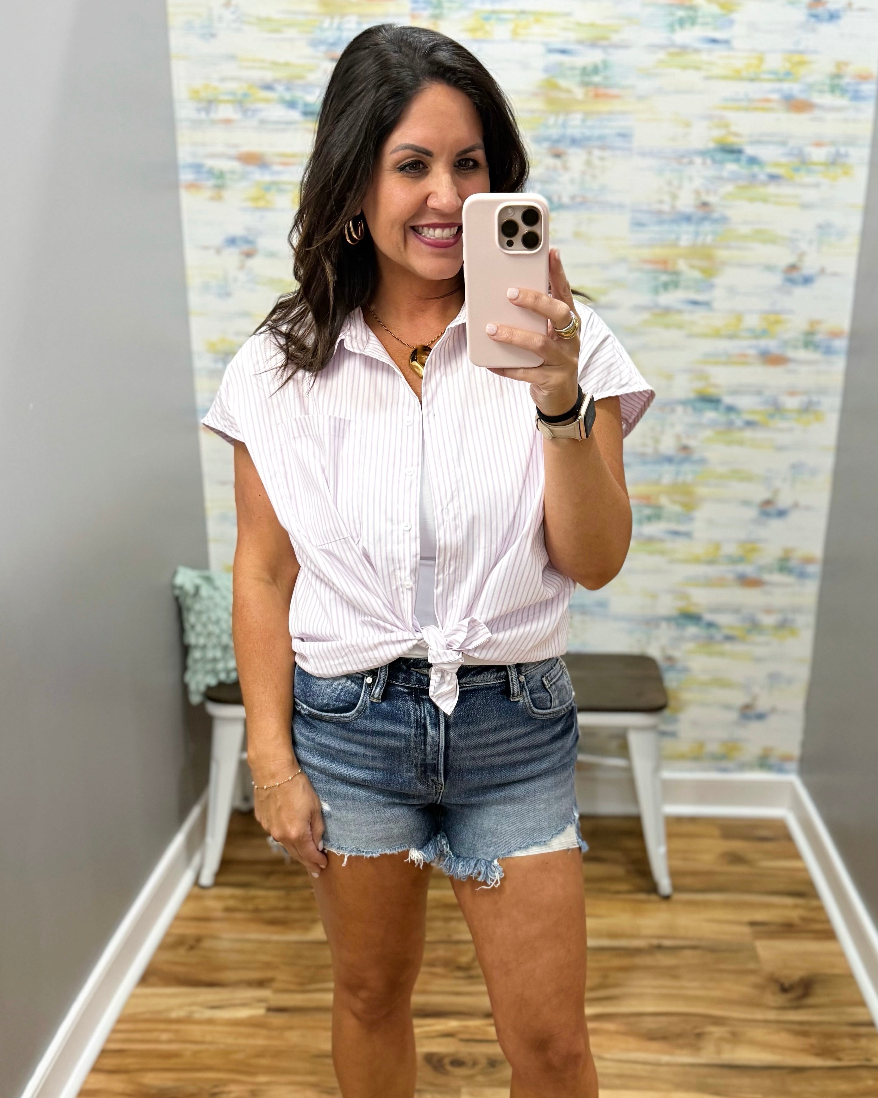 Woman taking a mirror selfie wearing a white striped shirt and denim shorts in a room with a colorful wall.