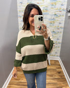 A woman takes a mirror selfie wearing a green and cream striped knit sweater, blue jeans, and gold jewelry in a home setting with wood floors and gray walls.