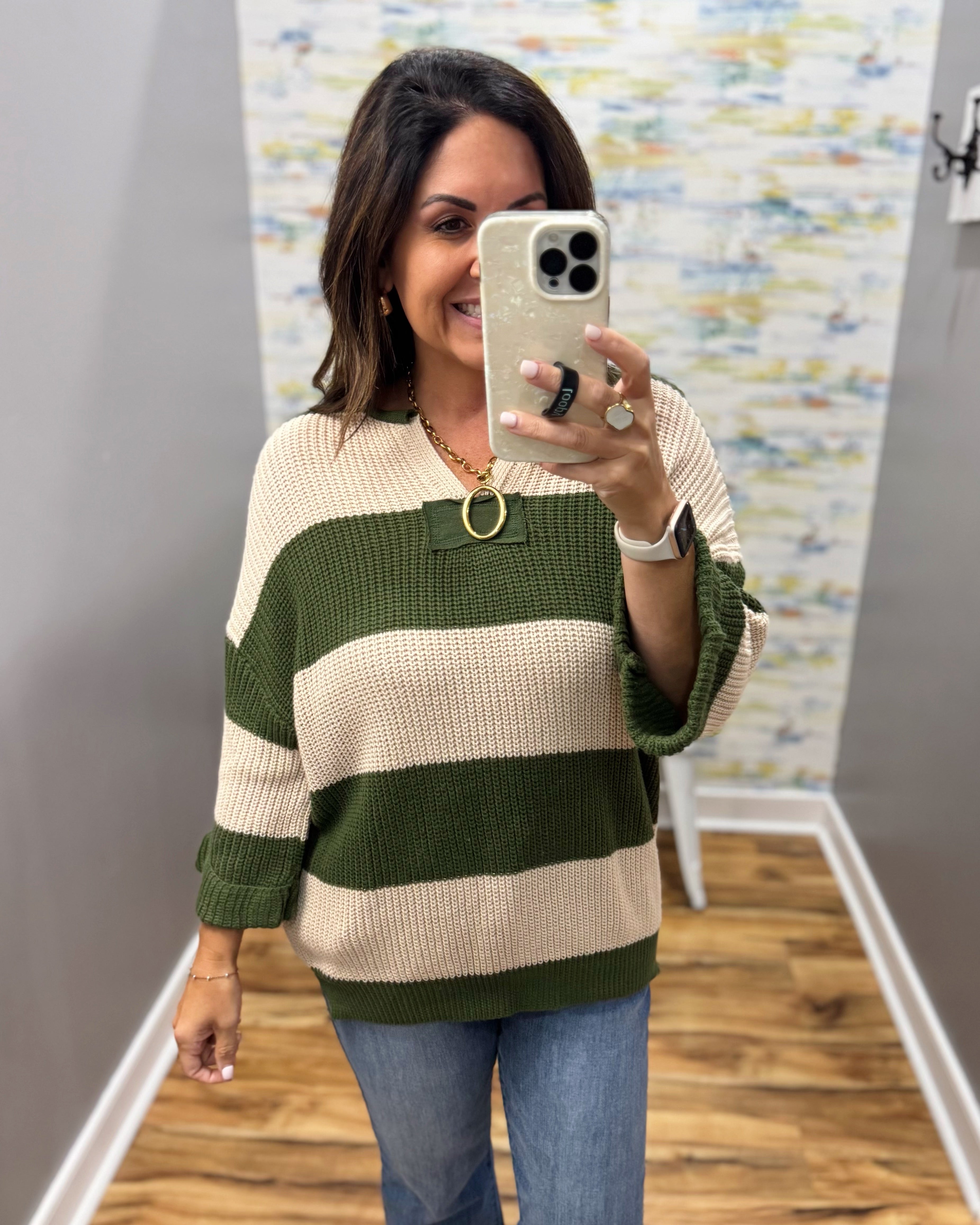 A woman takes a mirror selfie wearing a green and cream striped knit sweater, blue jeans, and gold jewelry in a home setting with wood floors and gray walls.