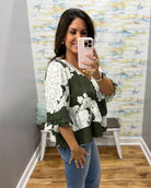 A woman takes a mirror selfie wearing a green and white floral print blouse with ruffled sleeves, paired with blue jeans, in an indoor setting with a patterned wallpaper background.