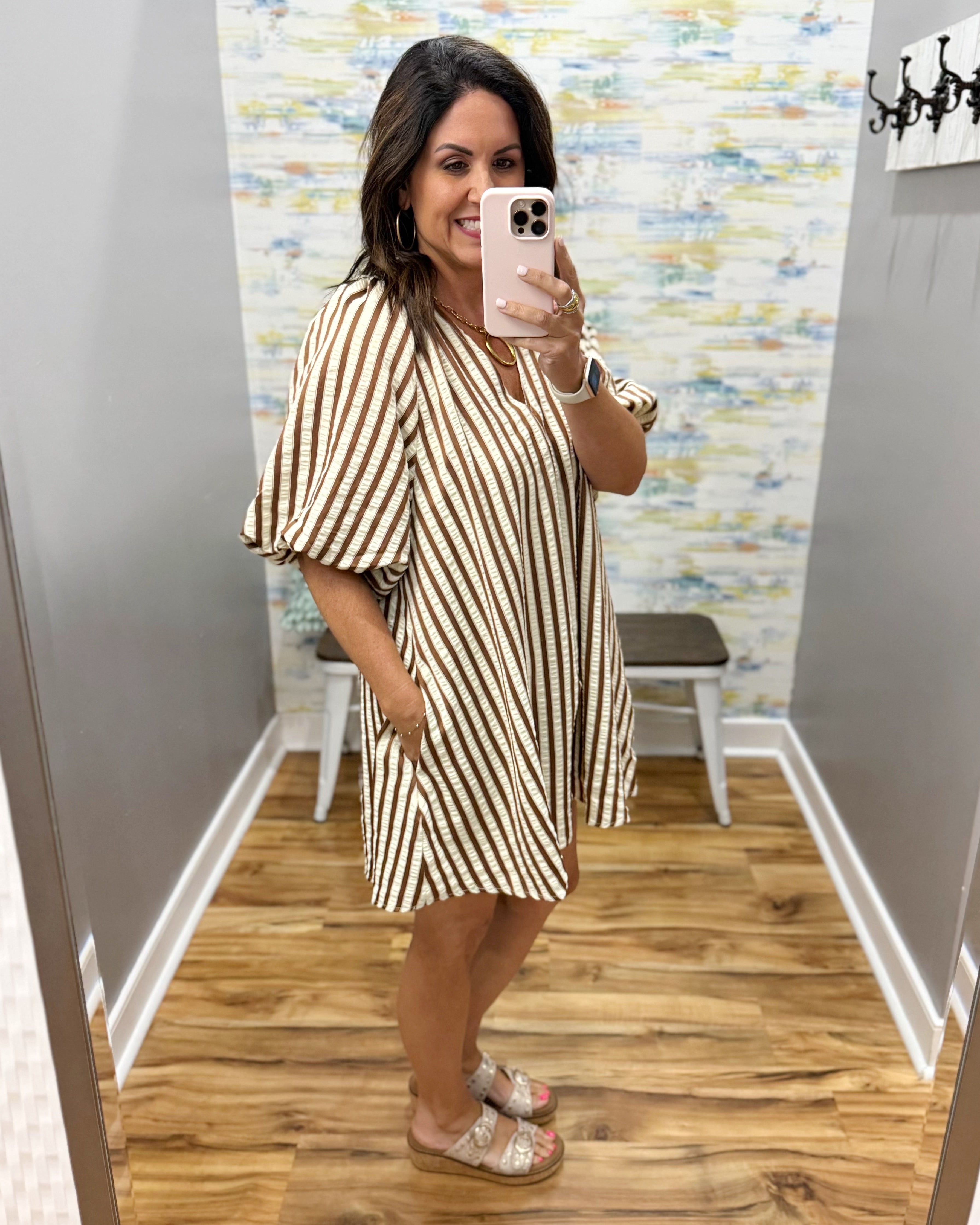 A woman takes a mirror selfie in a fitting room wearing a beige and brown vertically striped short-sleeve tunic dress with a V-neckline.