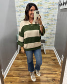 A woman takes a mirror selfie in a fitting room wearing a waffle-knit sweater with horizontal stripes in dark green and cream, paired with blue jeans and white sneakers.