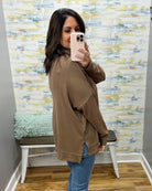 A woman takes a mirror selfie in a fitting room wearing a brown oversized long-sleeve top and blue jeans, with a patterned wall and a bench in the background.