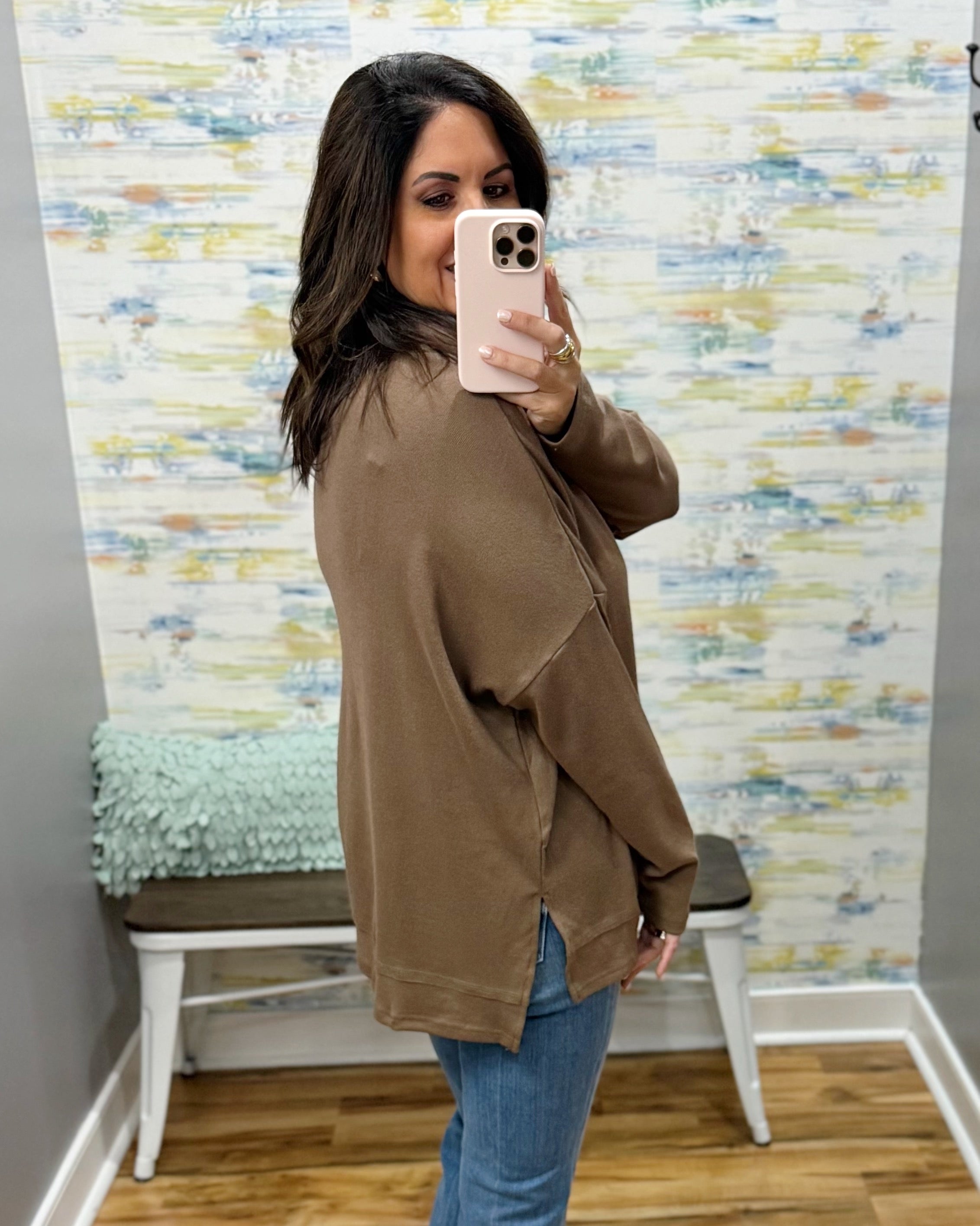 A woman takes a mirror selfie in a fitting room wearing a brown oversized long-sleeve top and blue jeans, with a patterned wall and a bench in the background.