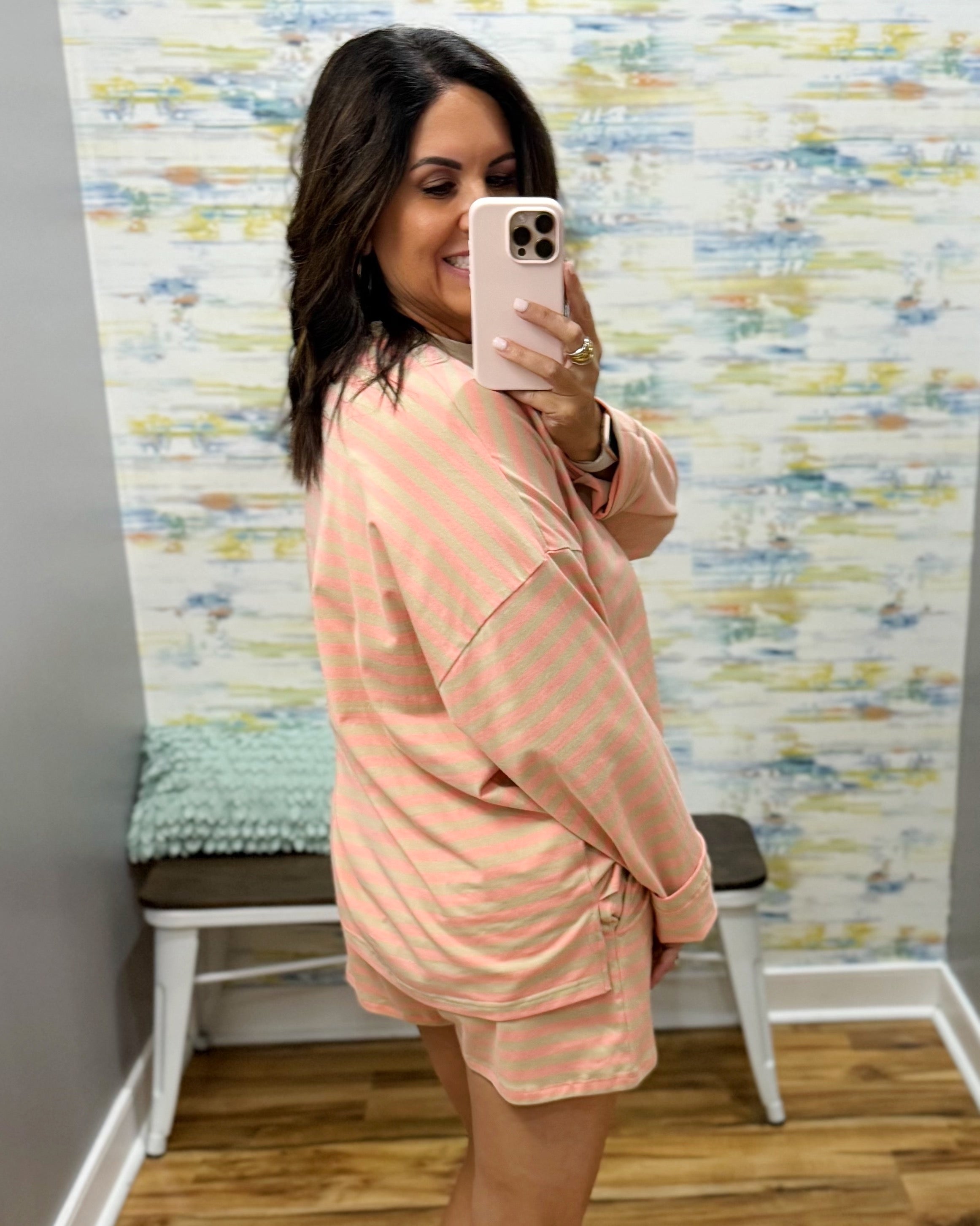 A woman takes a mirror selfie wearing a peach and white striped oversized long-sleeve top and matching shorts in a fitting room with a patterned wallpaper background.