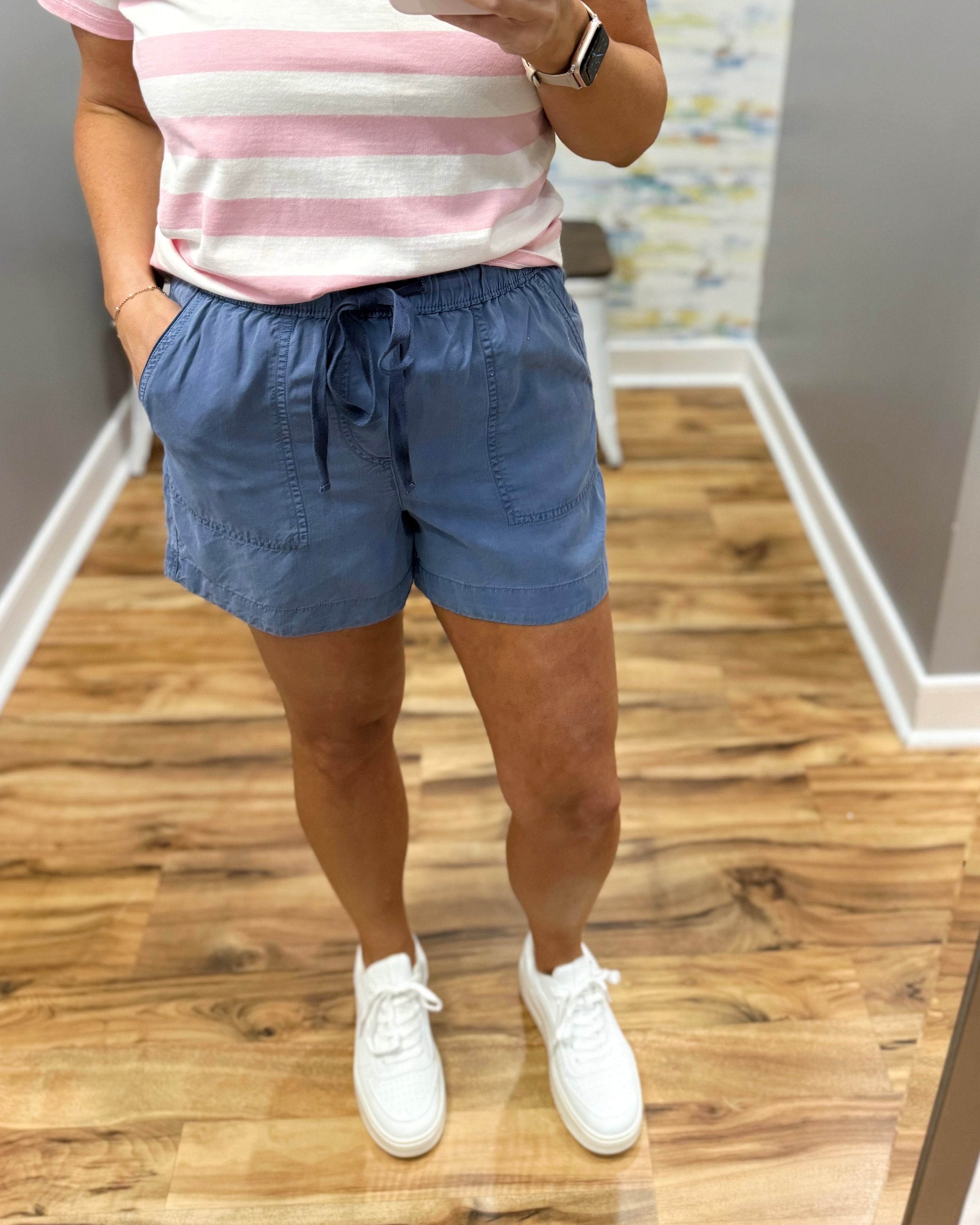 Person wearing a striped shirt, blue shorts, and white sneakers on a wooden floor.