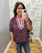 A woman taking a mirror selfie wearing a dark purple short-sleeve blouse with pink polka dots and a pink V-neck trim, paired with blue jeans.
