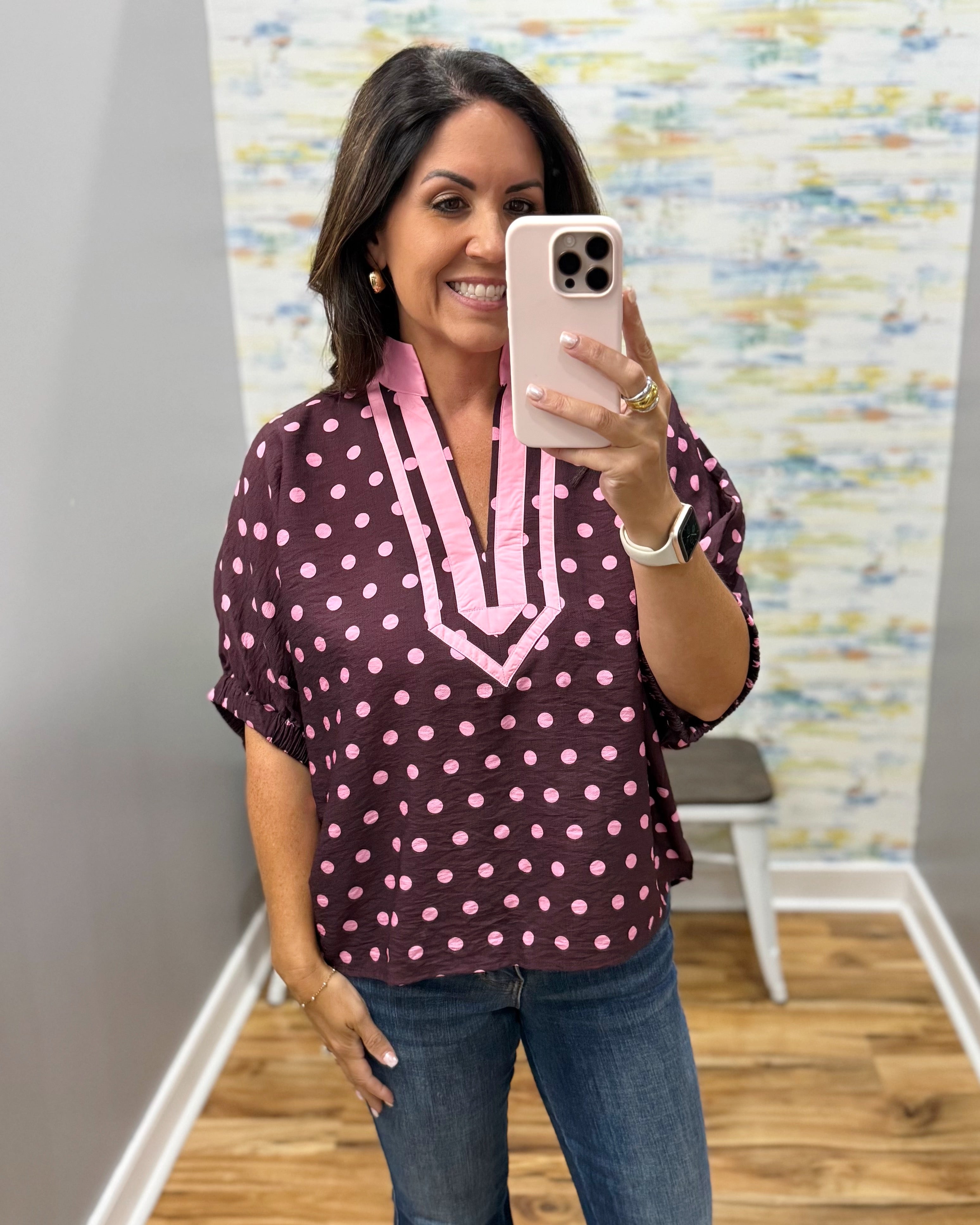 A woman taking a mirror selfie wearing a dark purple short-sleeve blouse with pink polka dots and a pink V-neck trim, paired with blue jeans.