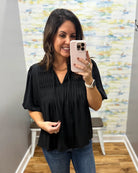 A woman takes a mirror selfie wearing a black short-sleeve blouse with pleated detailing and blue jeans in a boutique setting.
