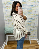 A woman takes a mirror selfie in a fitting room wearing a cream-colored, long-sleeve button-up shirt with dark brown vertical stripes and blue jeans.